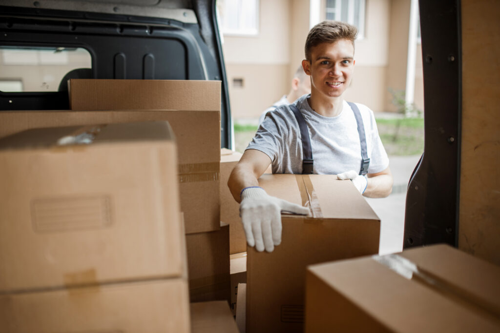 A young handsome smiling worker wearing uniform is standing next to the van full of boxes. House move, mover service